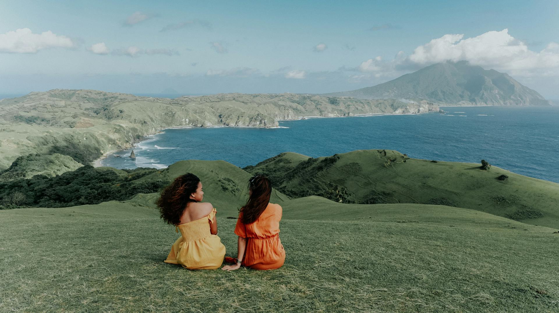 Back View of Two Girls Sitting on Grass Field in Front on Mountains