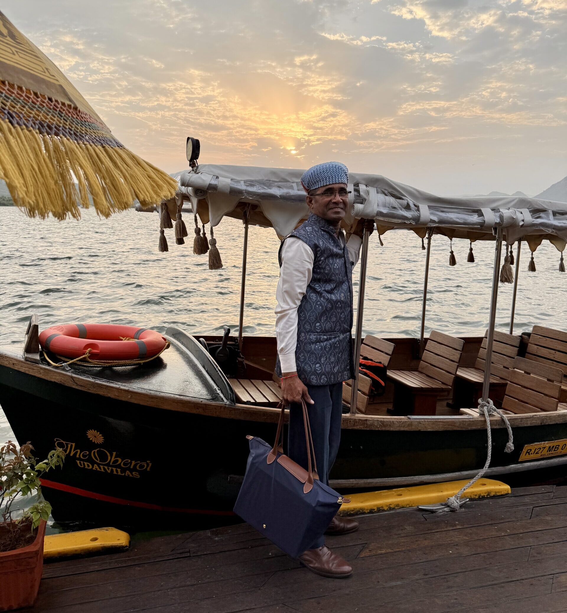 An Oberoi Udaivilas staff member in a printed blue vest and turban carries a guest’s bag beside the hotel’s canopied boat at the dock, with the sun setting over Lake Pichola in Udaipur