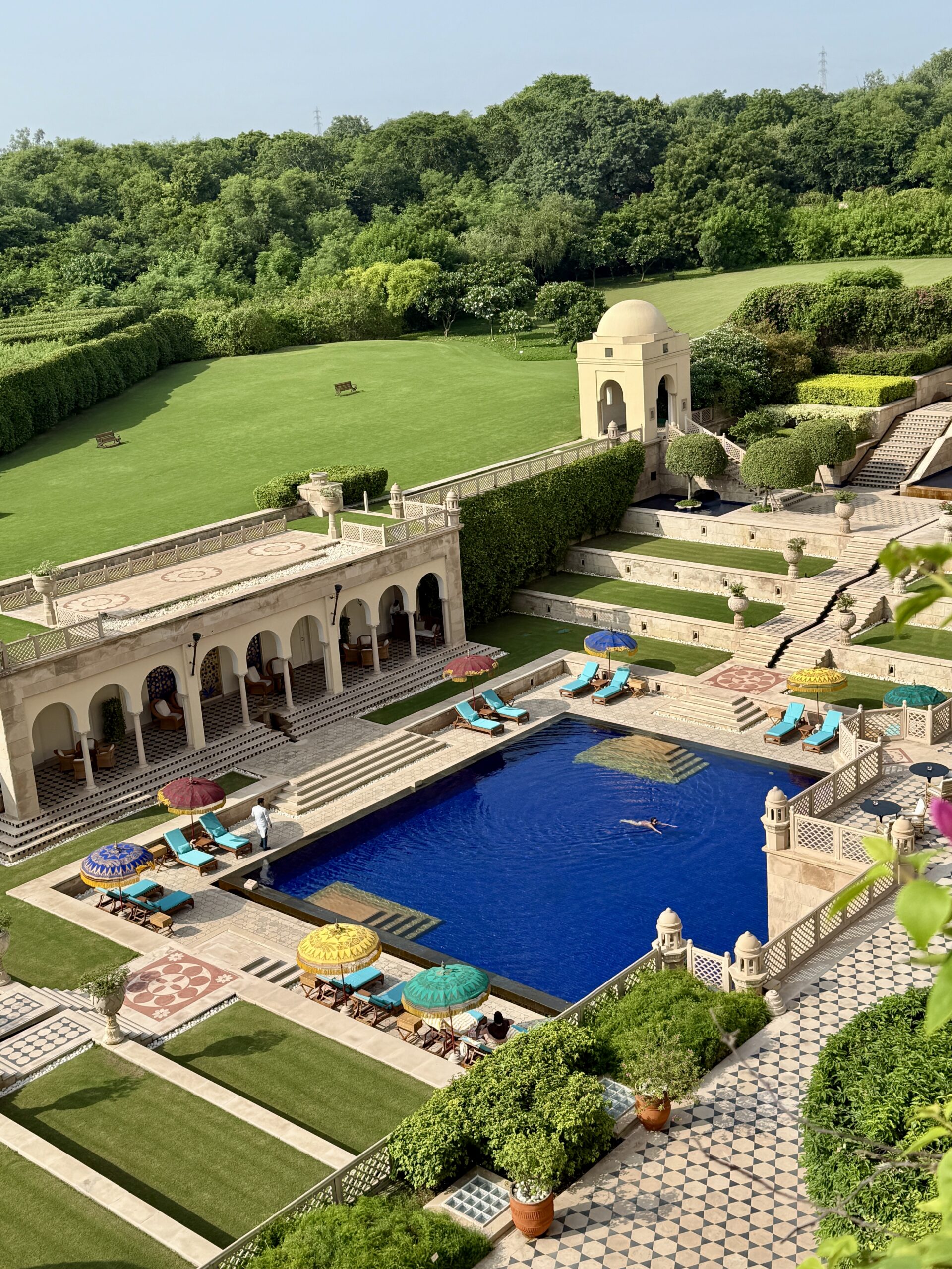 Overhead view of the terraced gardens at The Oberoi Amarvilas, Agra, with a deep blue swimming pool ringed by colorful parasols and turquoise loungers, arched pavilions and green lawns