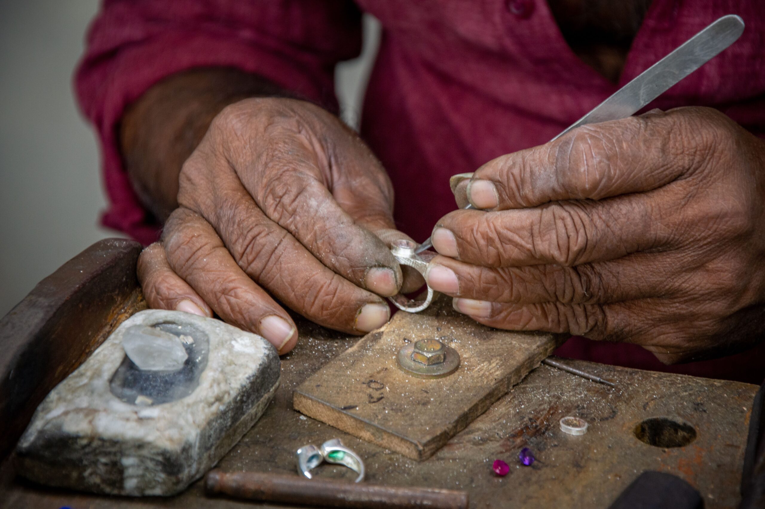 Close-up of a jeweler’s weathered hands setting a stone in a silver ring at a wooden workbench, with loose pink and purple gemstones and finished rings beside the tools