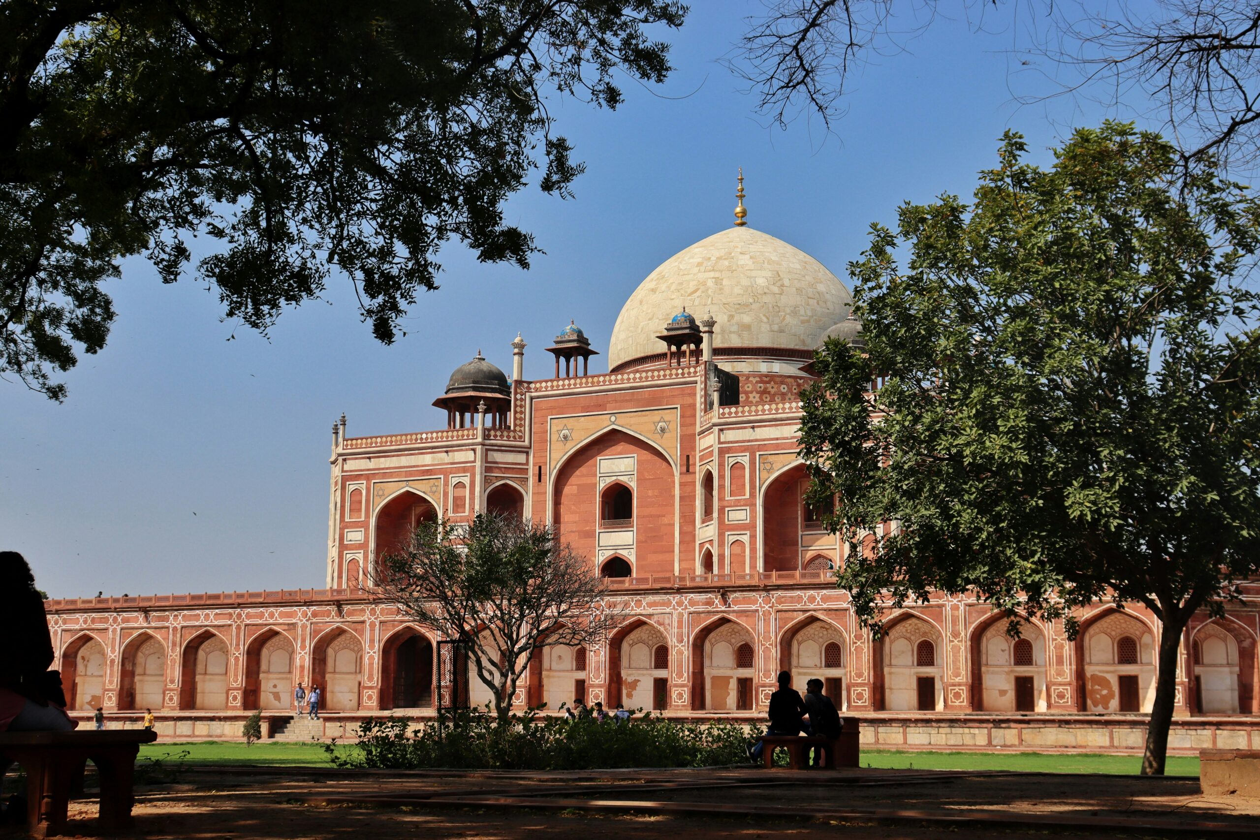 Humayun’s Tomb in Delhi, a red sandstone mausoleum with a white marble dome and rooftop chhatris, framed by shade trees with visitors resting on benches in the garden